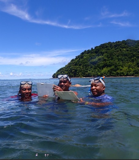 Photograph of three scuba divers in the sea. Their heads and shoulders are above the water and they are looking at screens, whilst undertaking a seagrass practical.