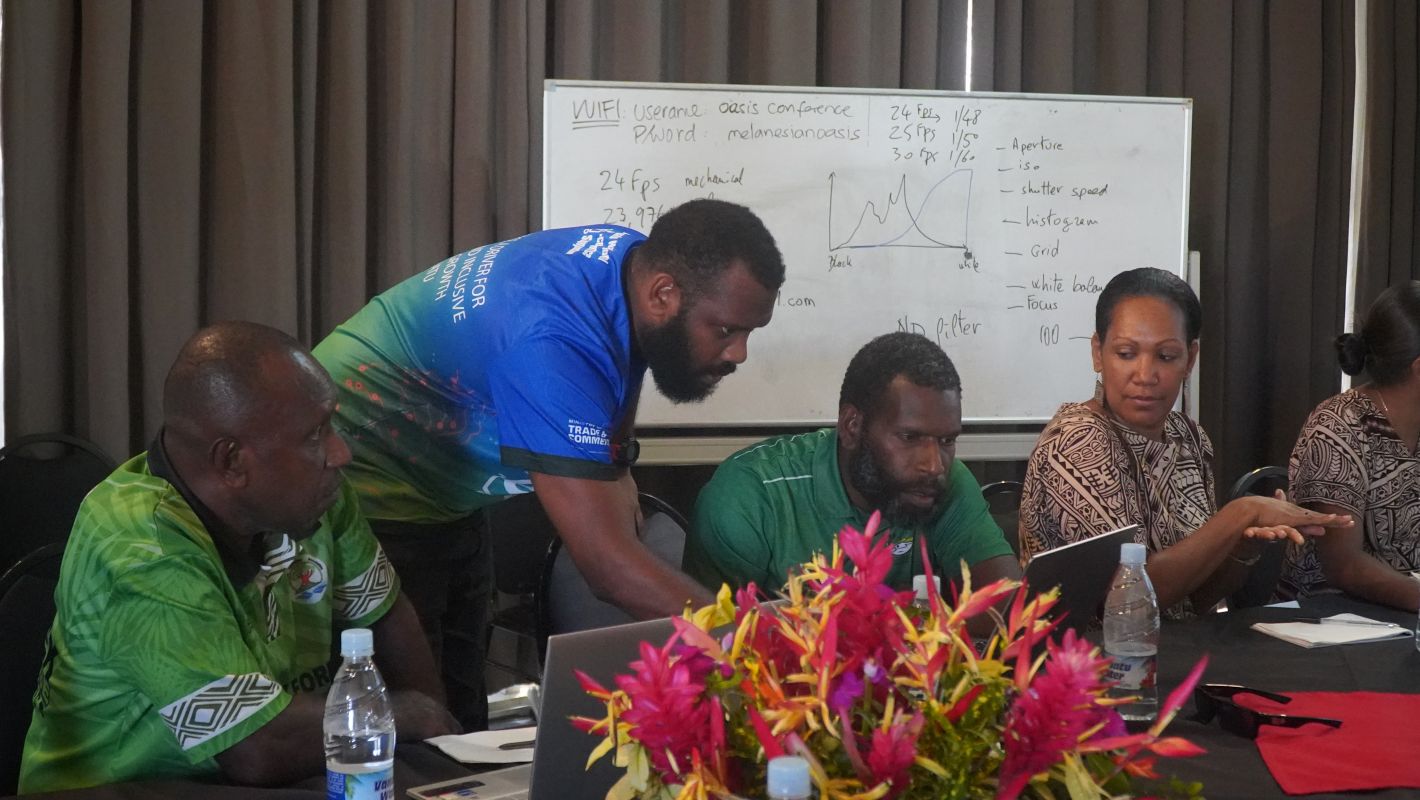 4 people sat along a table with another person leaning over showing them something on a laptop